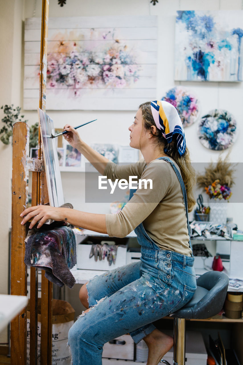 Side view of female artist sitting near easel and drawing colorful picture with paints while working in creative workshop