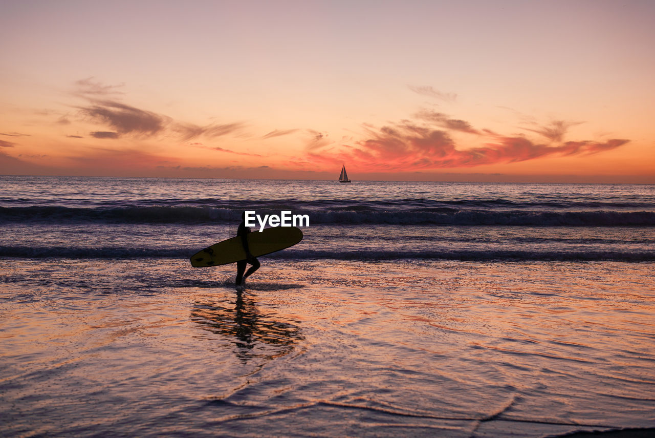 Side view of man with surfboard walking on shore at beach against sky during sunset