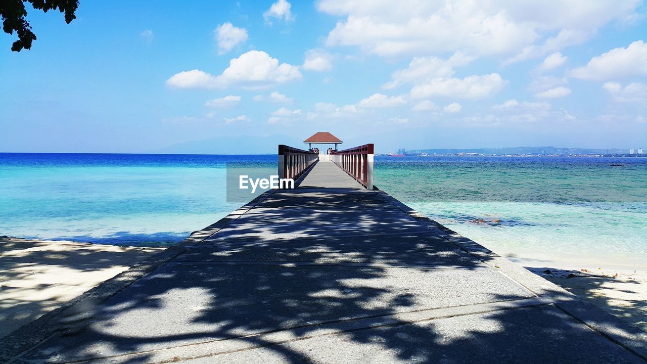 Pier leading towards gazebo at beach against cloudy sky during sunny day