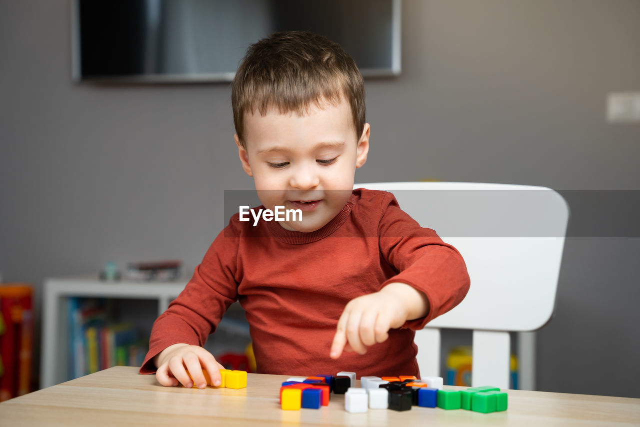 A cute happy little toddler boy of two years old sits at a children's table and plays 