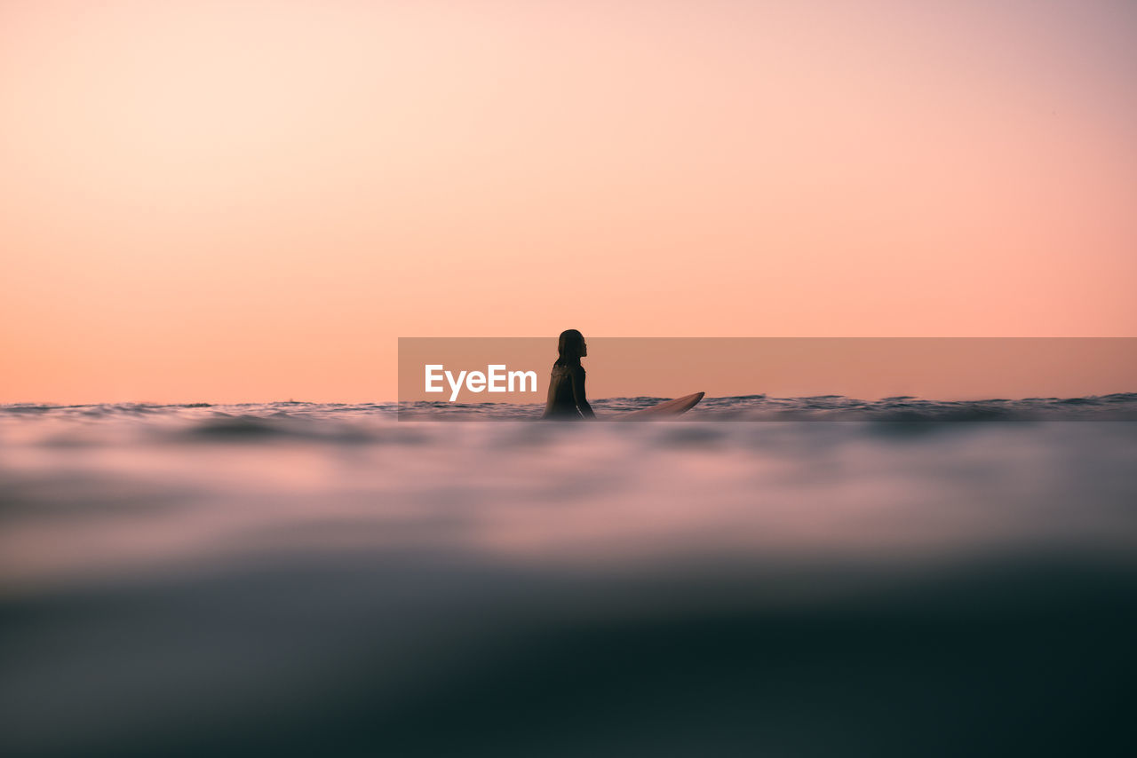 Rear view of woman sitting on a surfboard in the  ocean against an orange peach sky during sunset