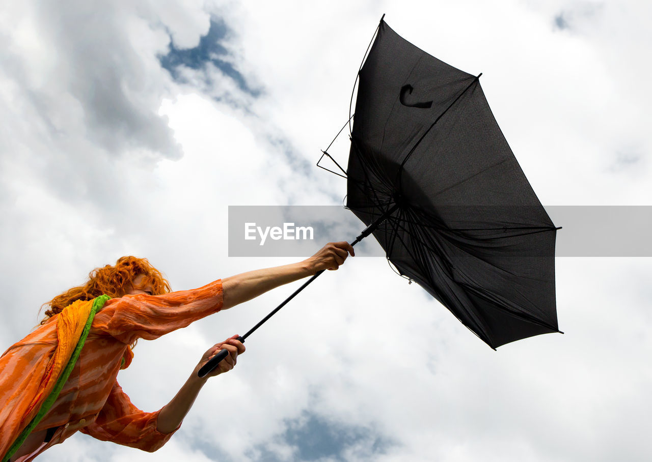 Low angle view of mature woman holding damaged umbrella against cloudy sky