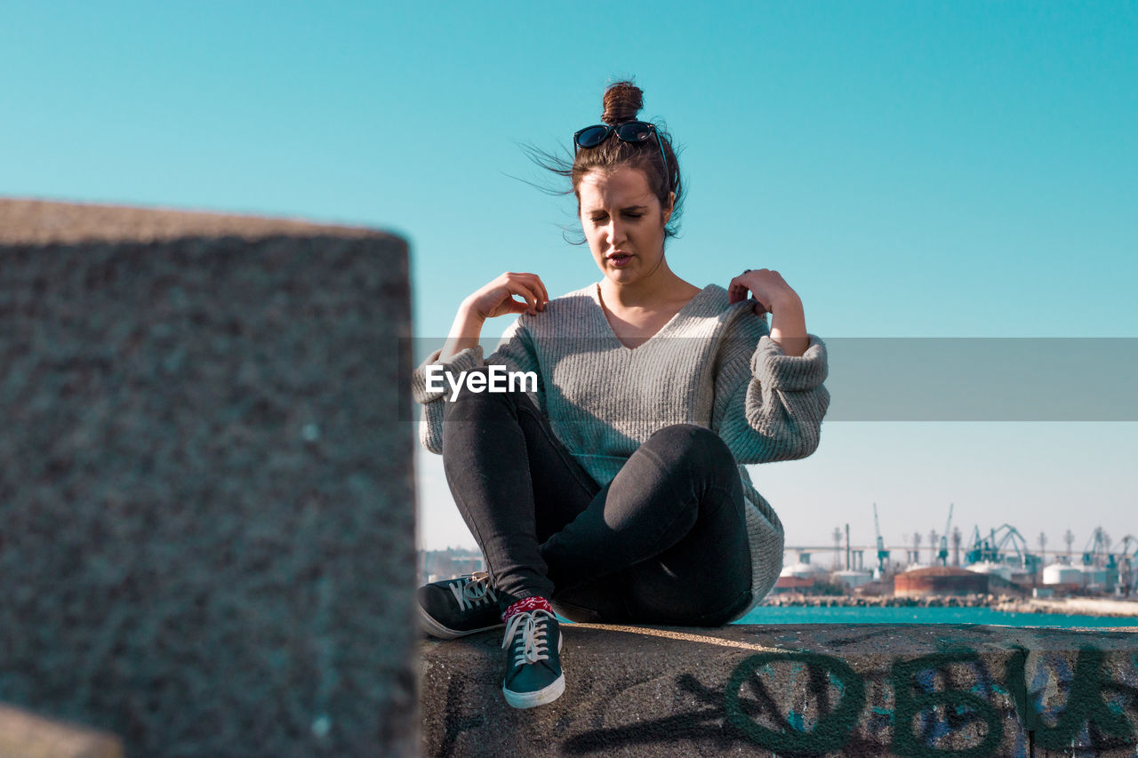 Young woman sitting on retaining wall against clear sky