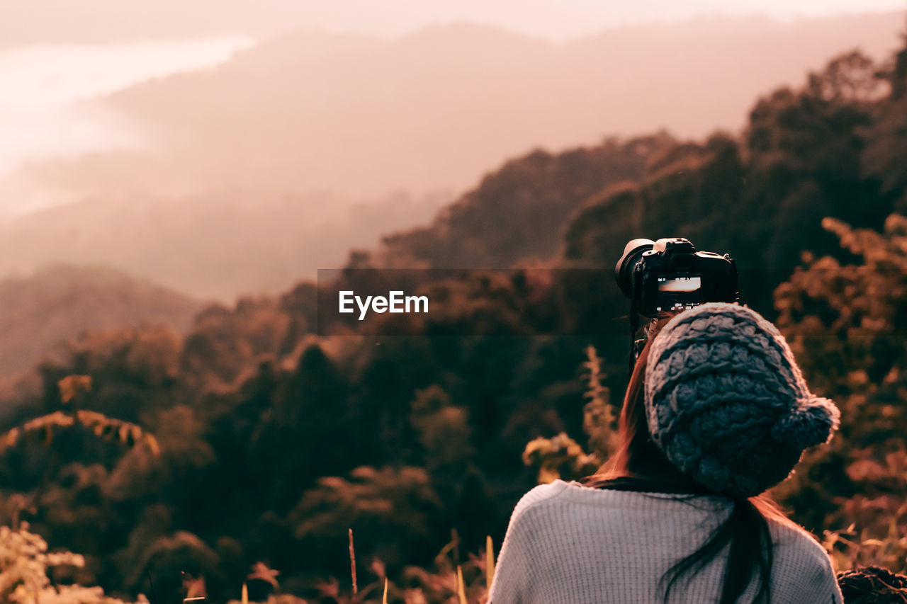 Rear view of mid adult woman photographing while standing on mountain against sky during sunset