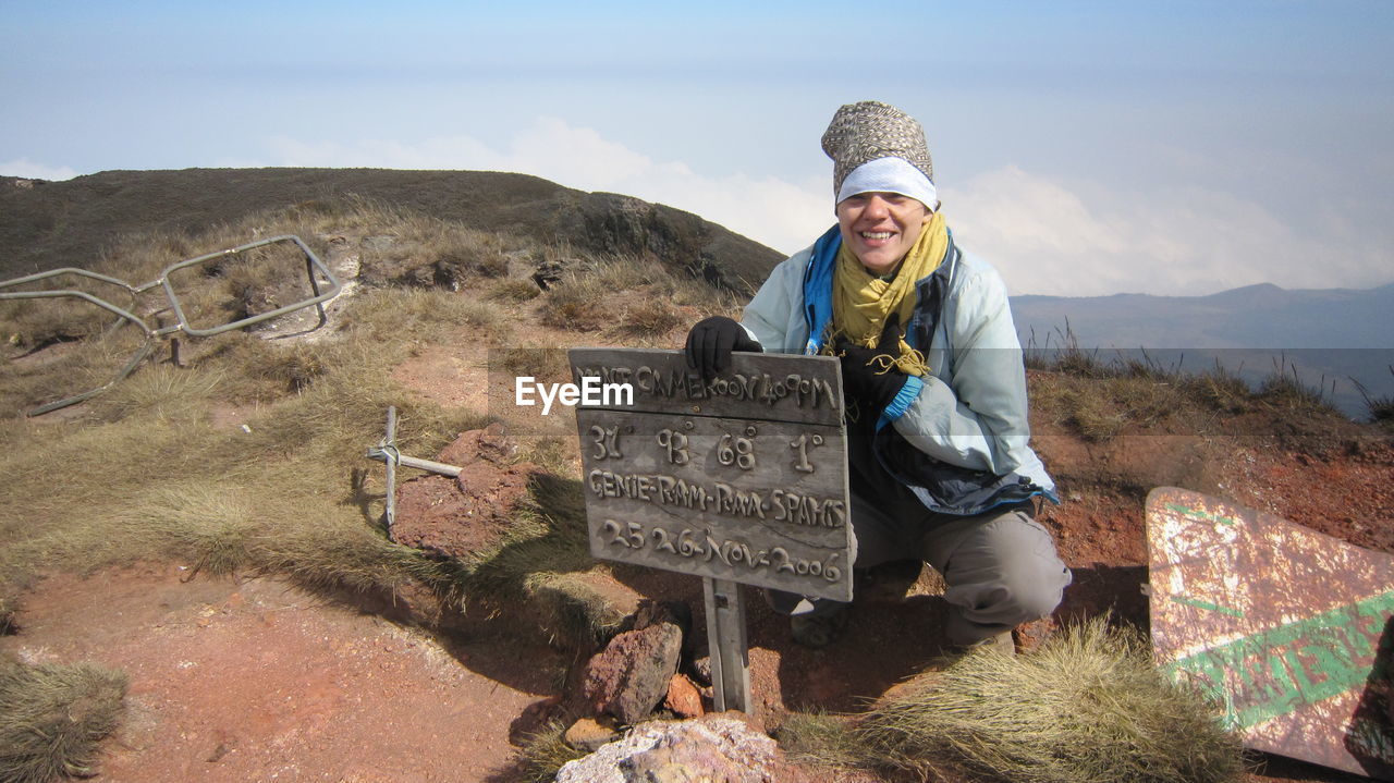 Smiling woman crouching by information sign against sky