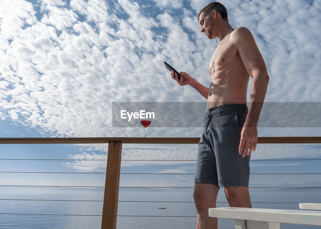 Low angle view of shirtless mature man using phone against cloudy sky