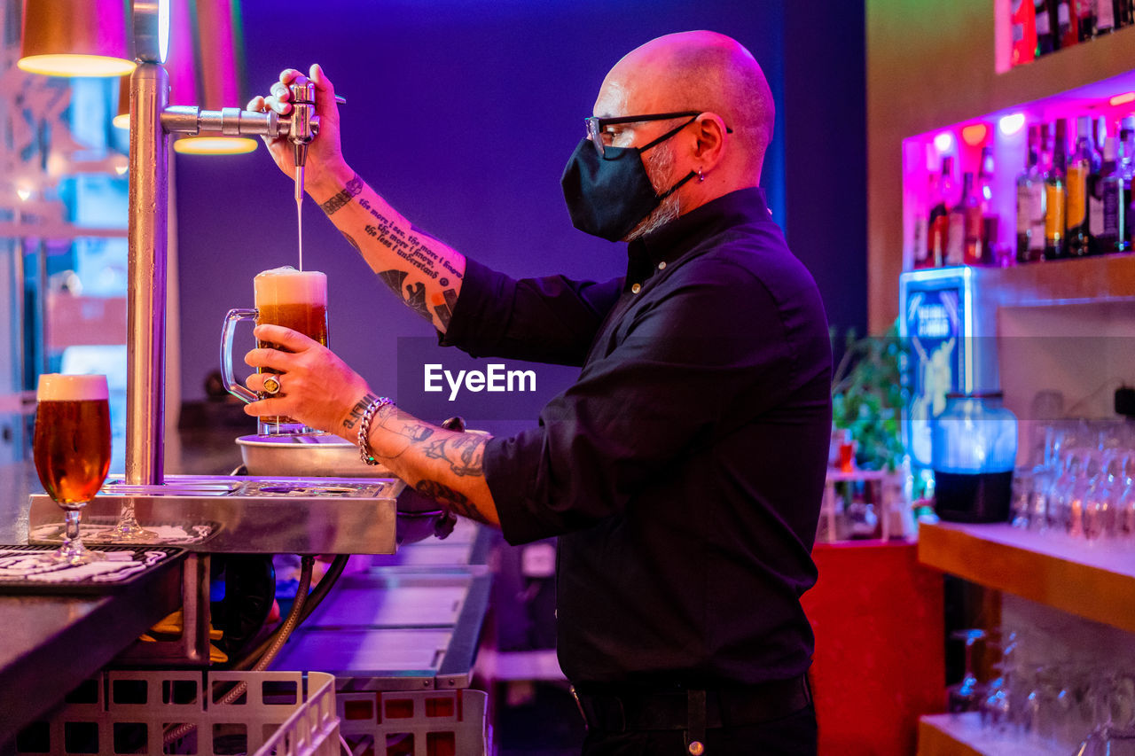 Side view of serious barkeeper in eyeglasses pouring beer into glass while working in bar during coronavirus pandemic