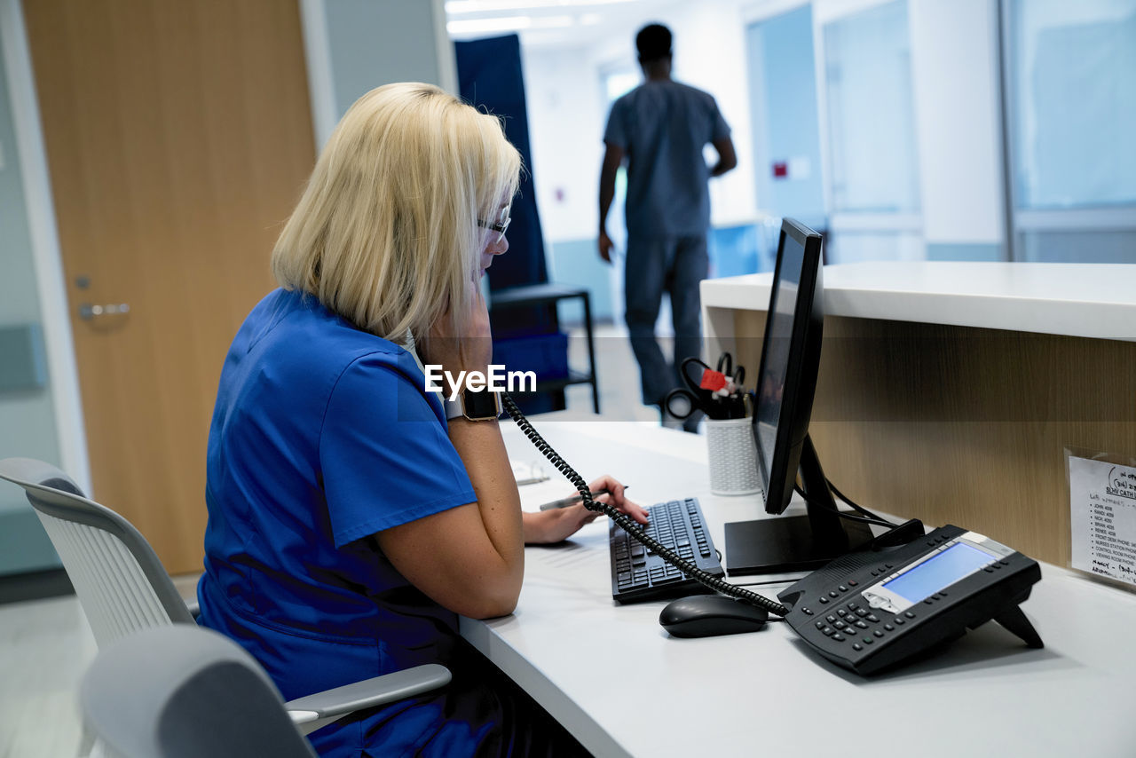 Female doctor talking on telephone while using desktop computer with male colleague in background