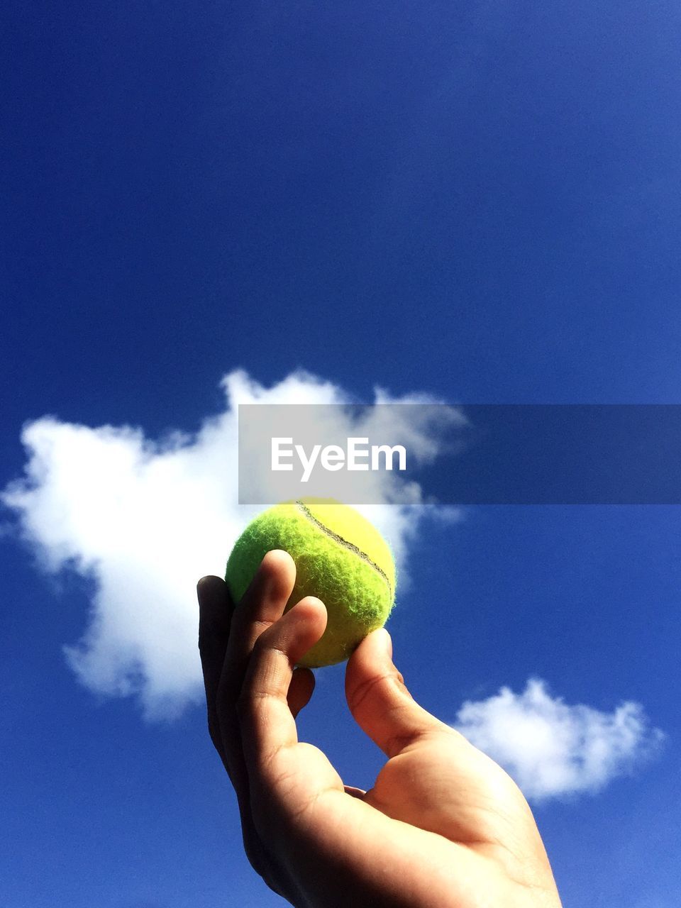 Cropped hand holding tennis ball against blue sky