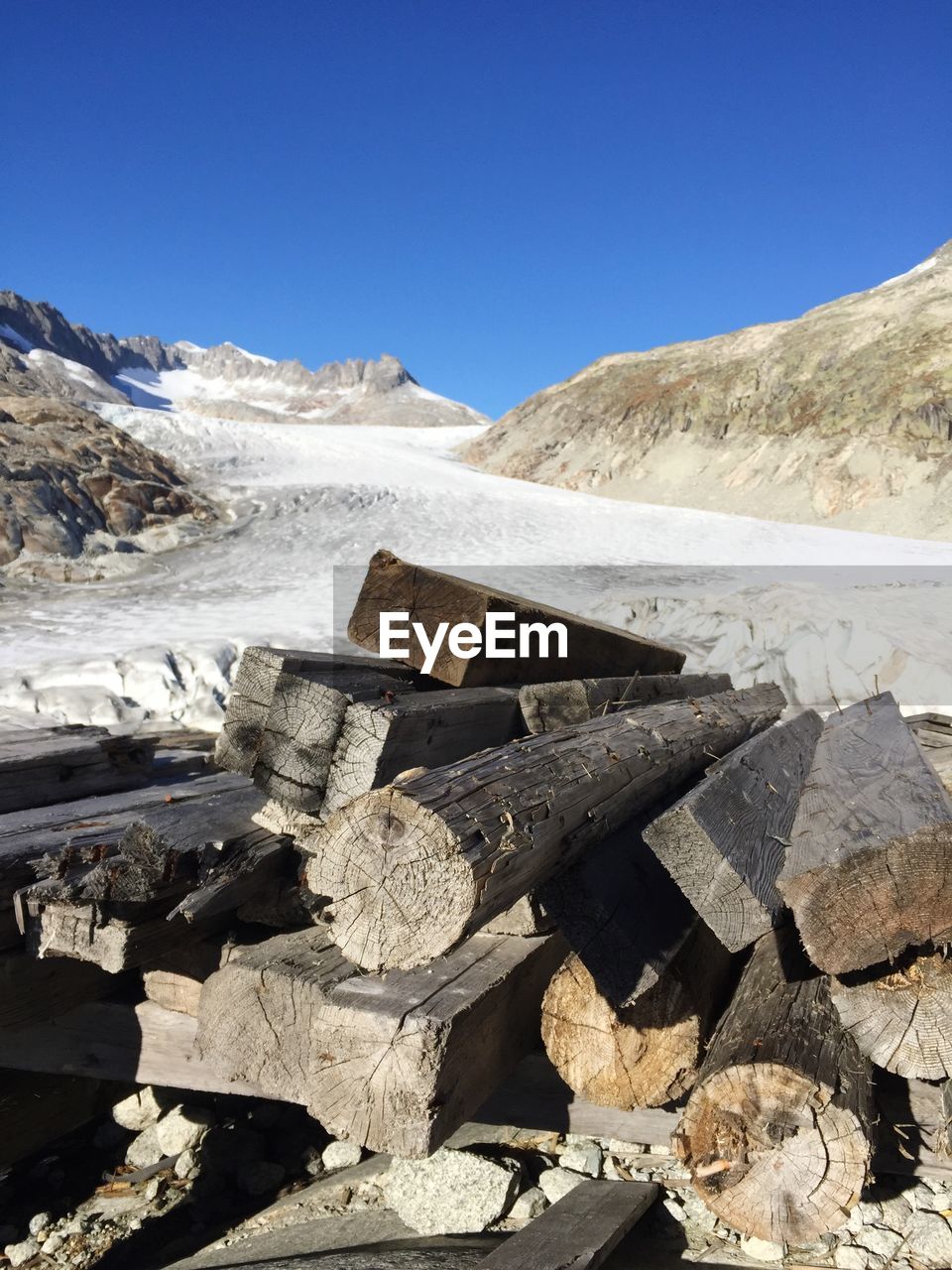 Stack of logs by snow covered mountains against clear blue sky