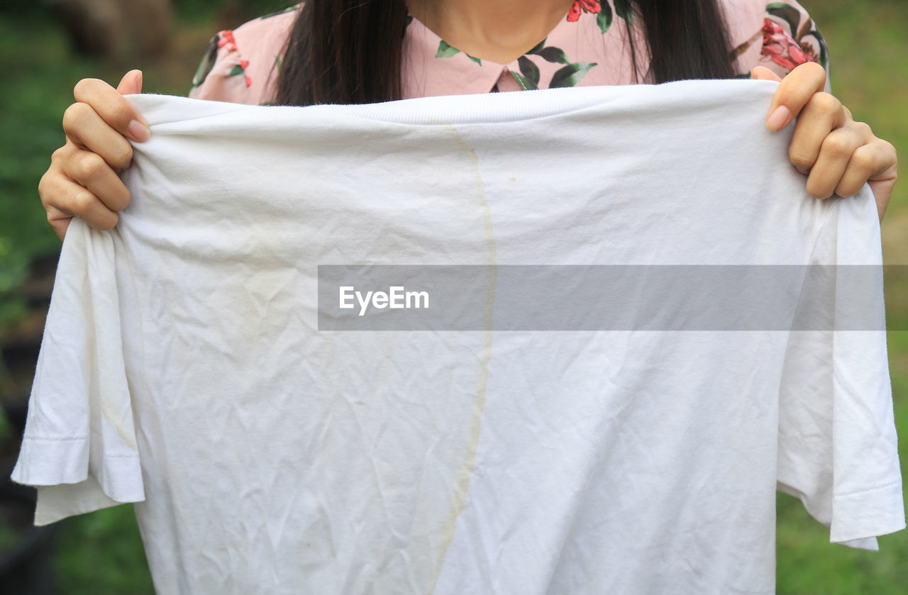 MIDSECTION OF WOMAN HOLDING WHITE DRESS STANDING AGAINST BLURRED BACKGROUND