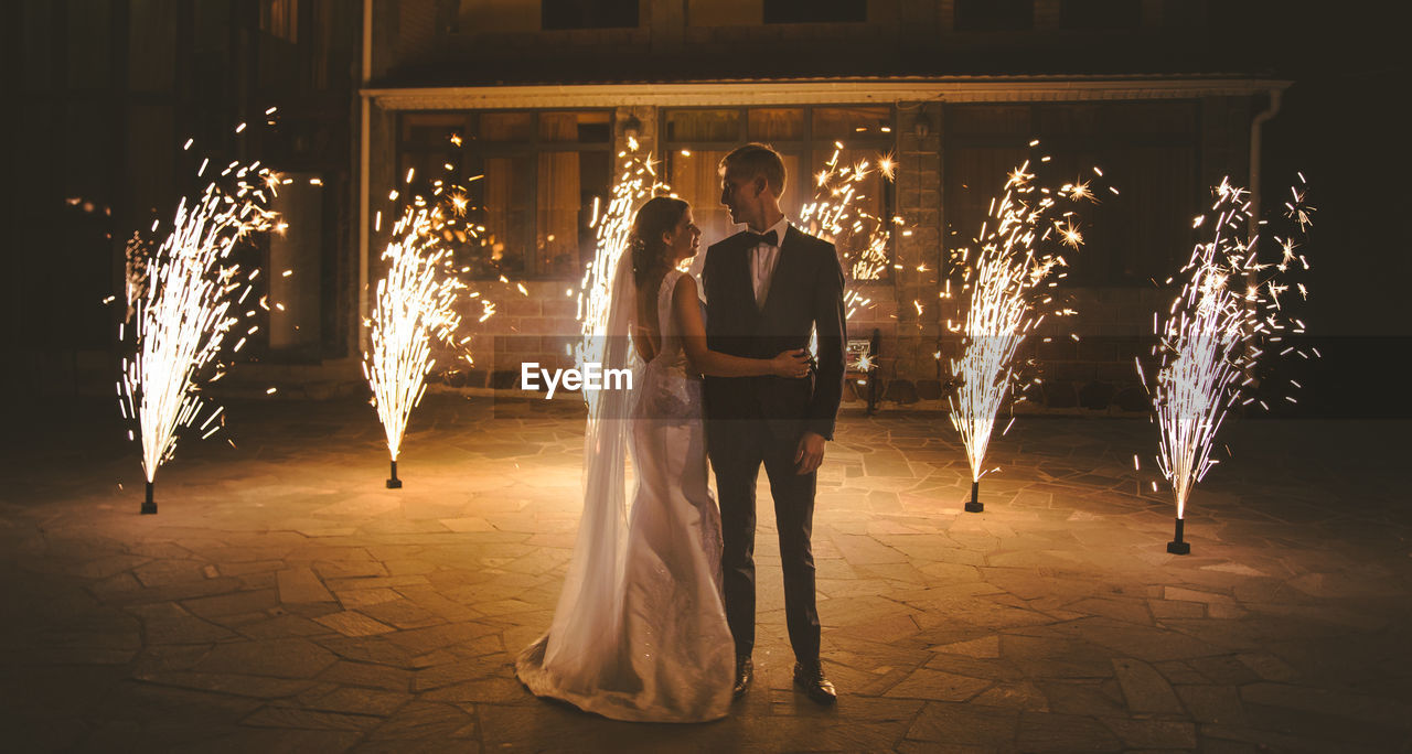 Bride and bridegroom standing against firework at night