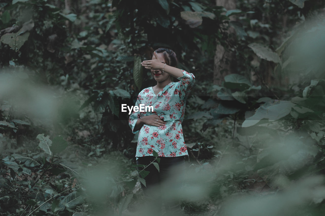 Smiling woman hiding eyes while standing in forest