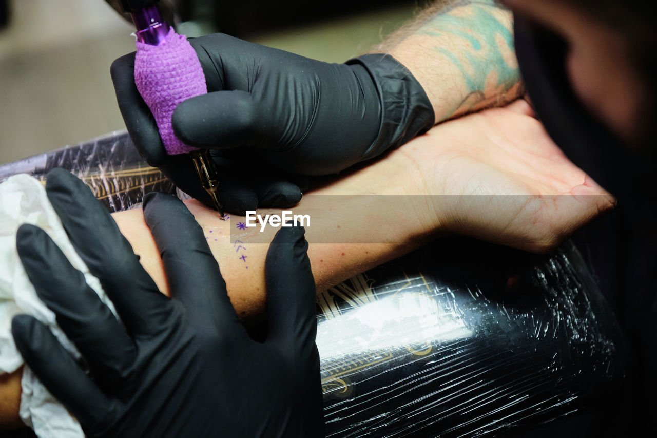 Unrecognizable concentrate bearded male master in black face mask and gloves creating tattoo on arm of crop faceless client in salon