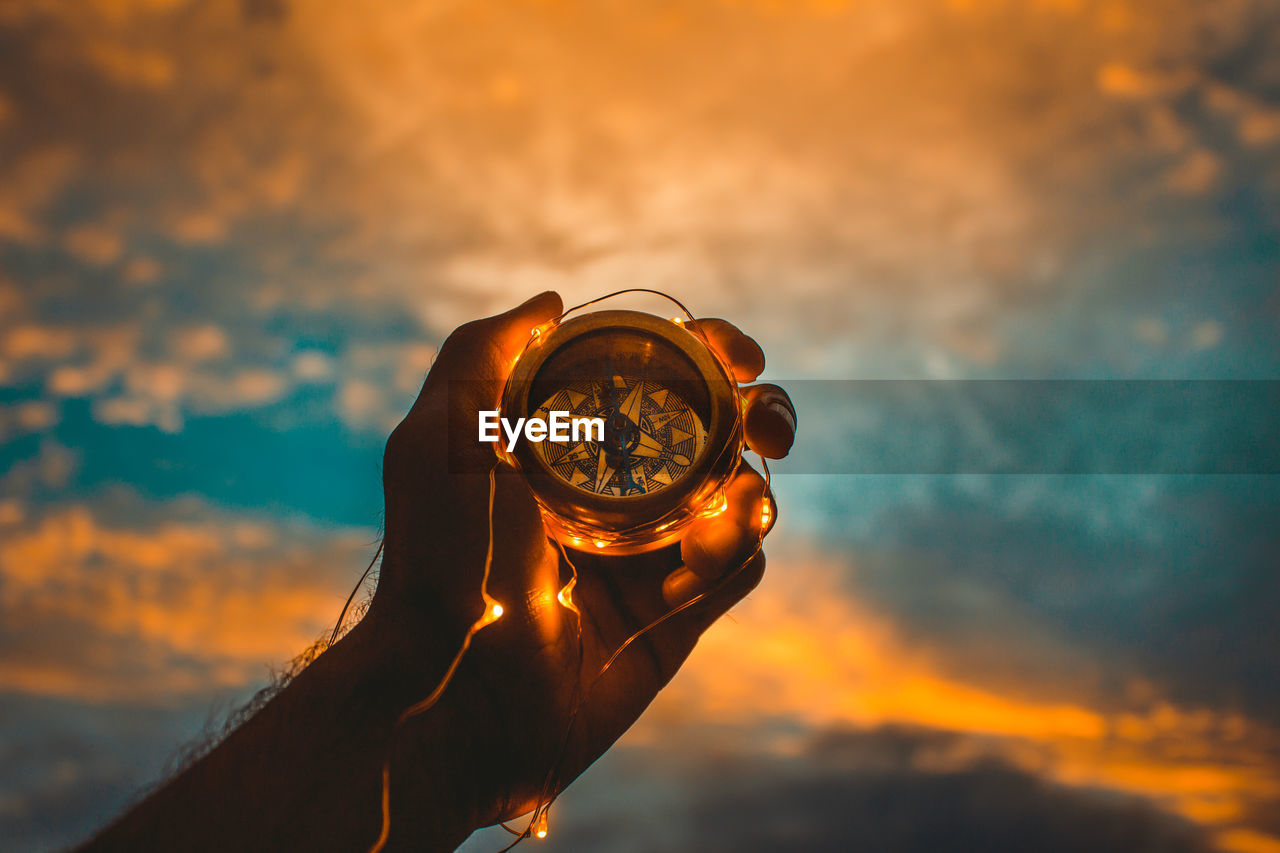 Cropped image of man holding illuminated string light and navigational compass against cloudy sky during sunset