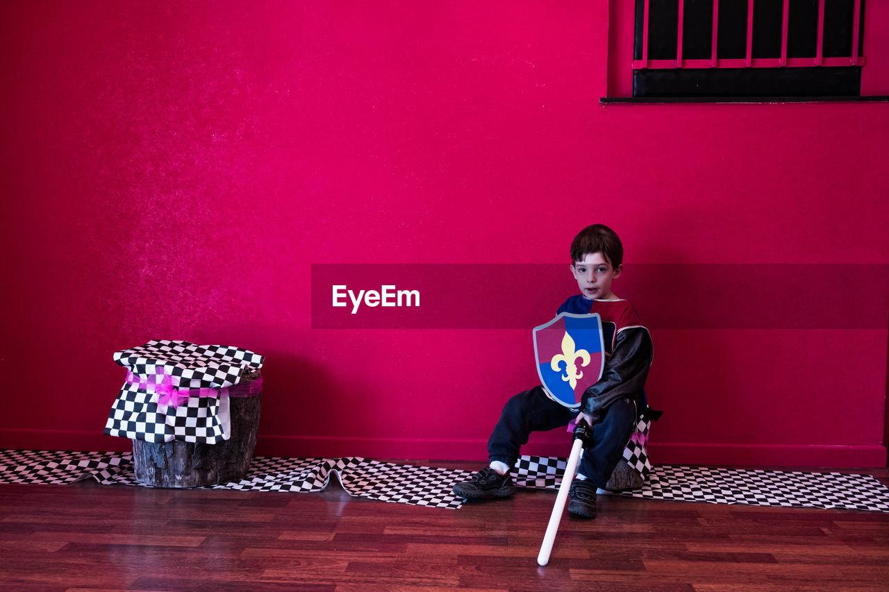 Full length portrait of boy in costume sitting against pink wall