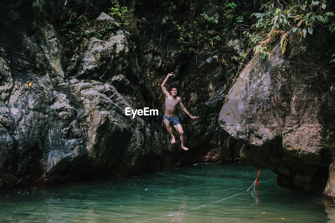 Full length of shirtless young man jumping in river by rock formations