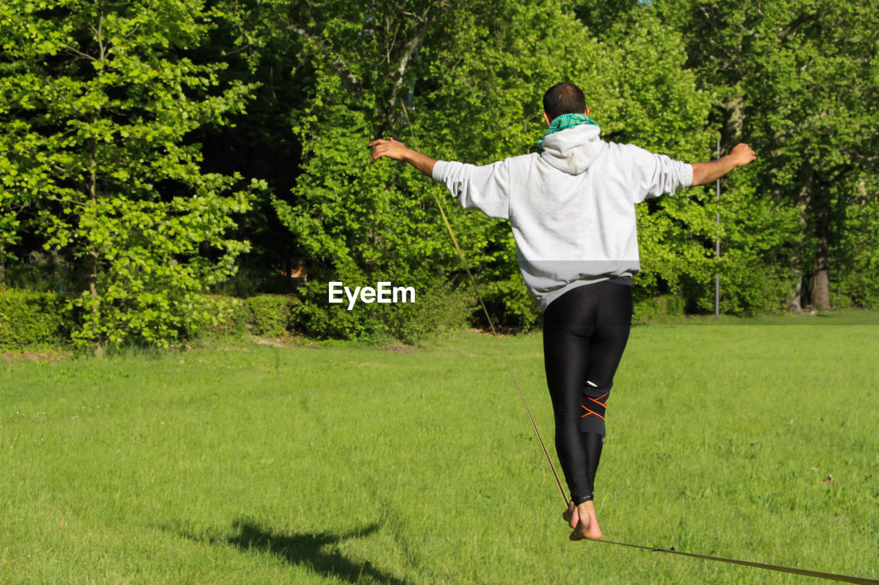 Rear view full length of man slacklining on grassy field during sunny day