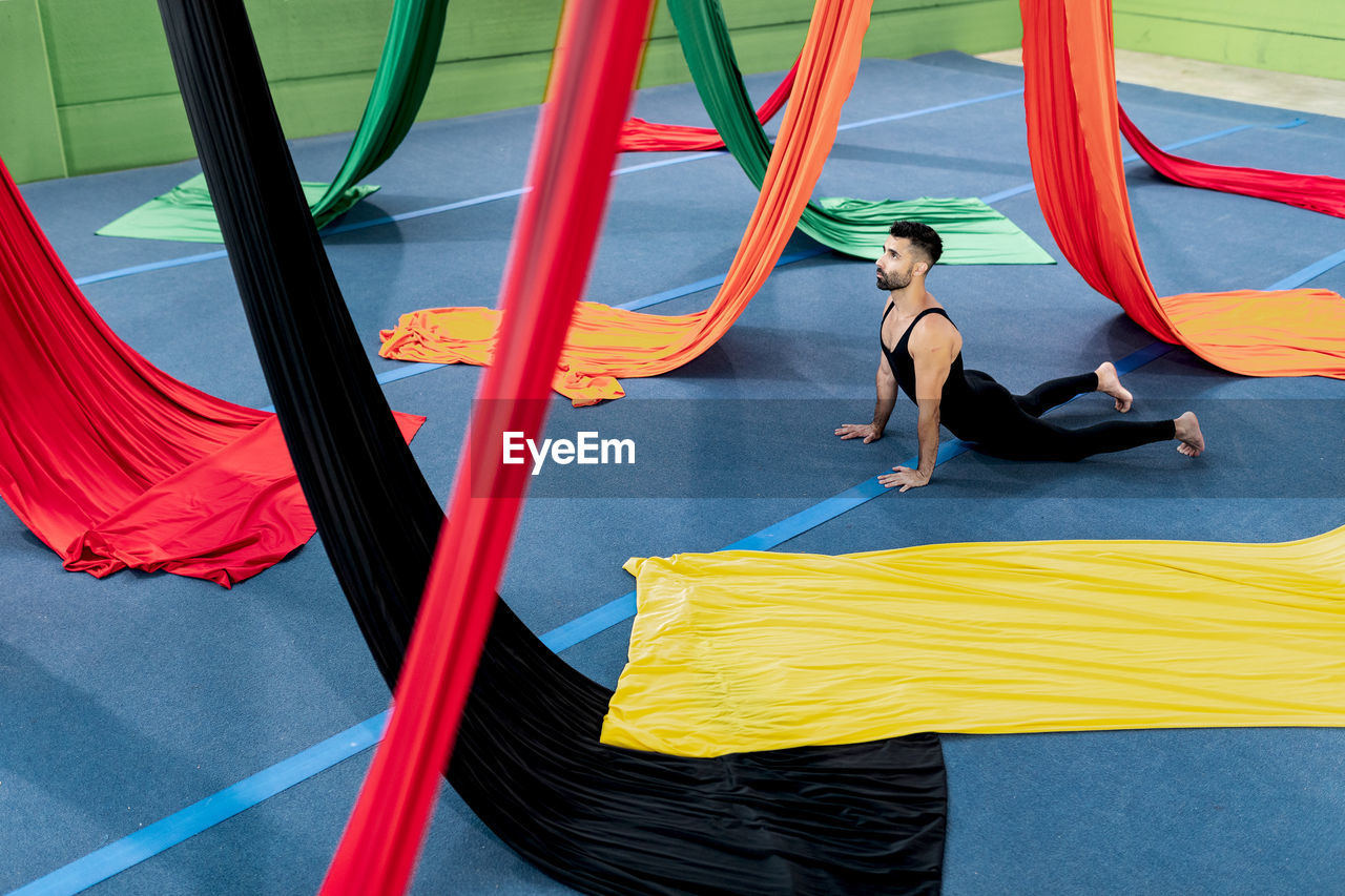 High angle of flexible man in leotard doing stretching exercise near colorful aerial silks during rehearsal in studio