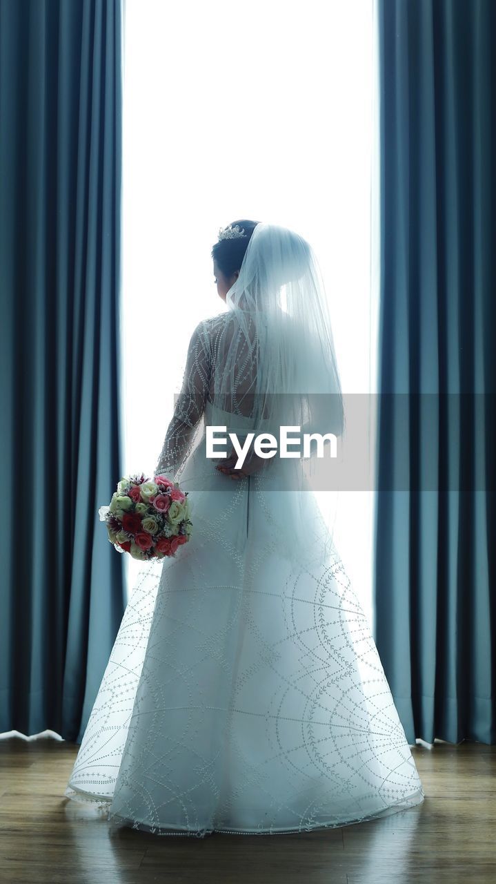 Rear view of bride wearing wedding dress standing with bouquet on hardwood floor
