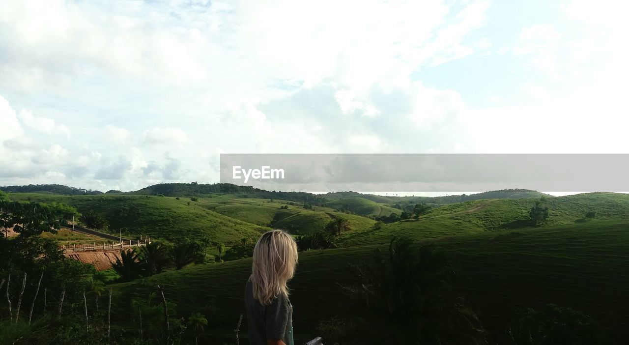 Side view of woman looking at grassy landscape against cloudy sky