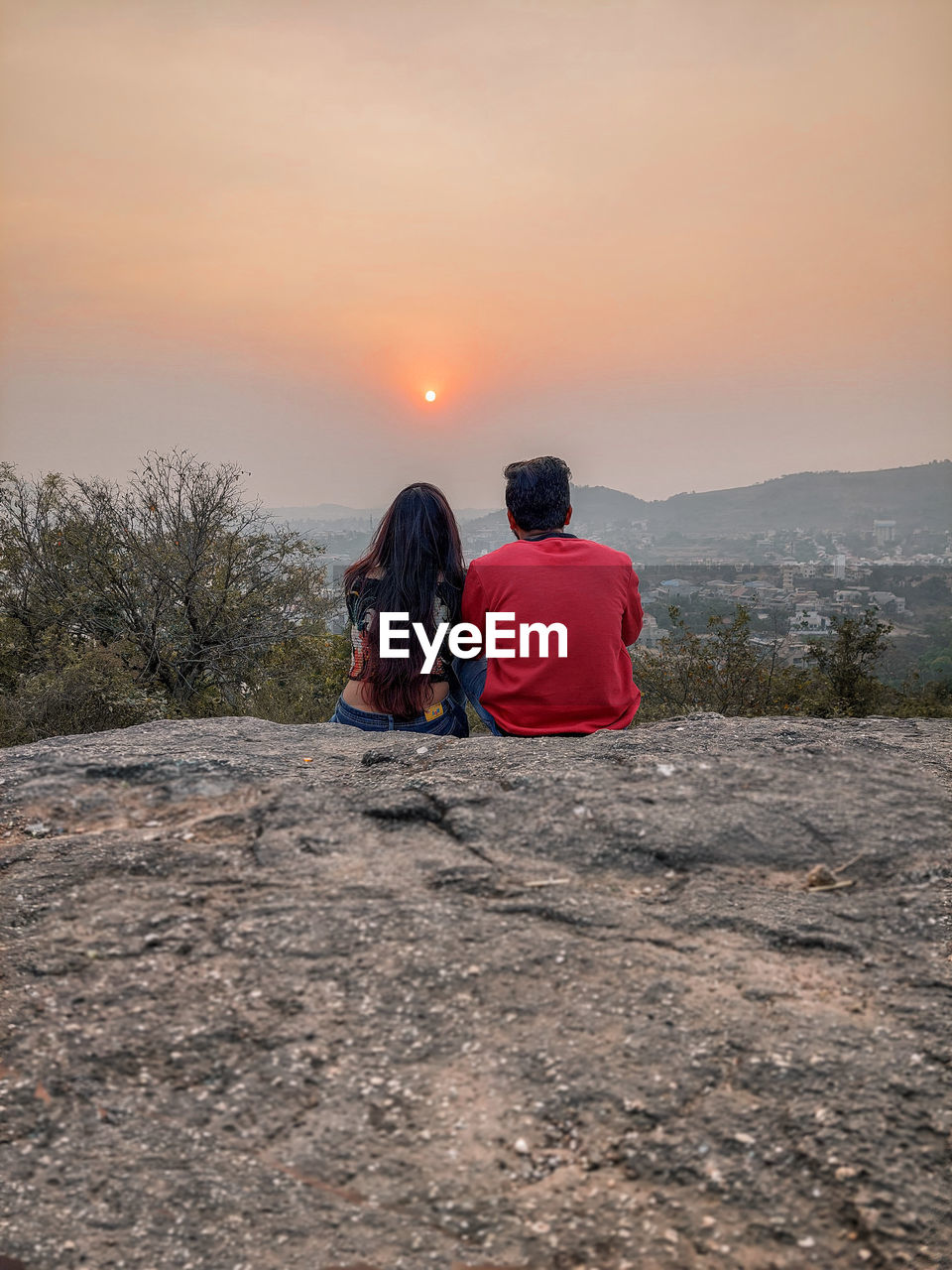 Rear view of couple sitting on edge of cliff gainst sunset