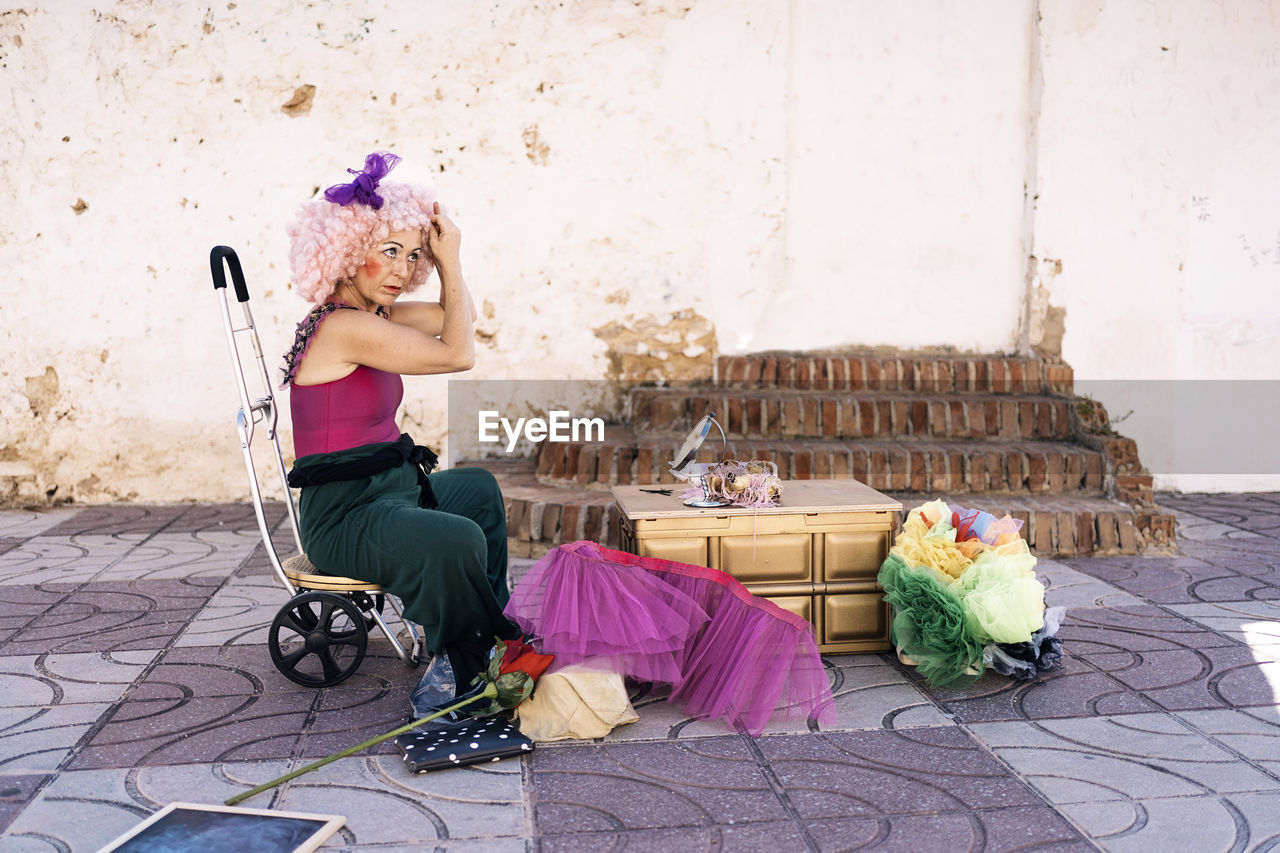 Side view of concentrate middle aged female clown with bright makeup in costume putting on wig and looking away while sitting on circus chair with wheels on street