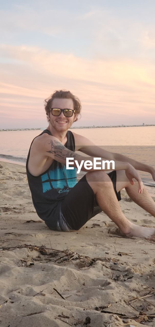 Portrait of man sitting on shore at beach against sky during sunset