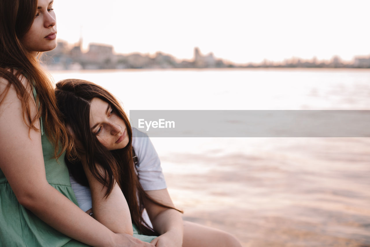 Thoughtful lesbian couple sitting by river in city at sunset