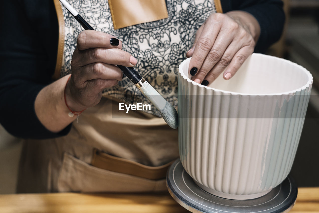 Crop anonymous female potter in apron painting handmade clay pot in professional workshop on blurred background