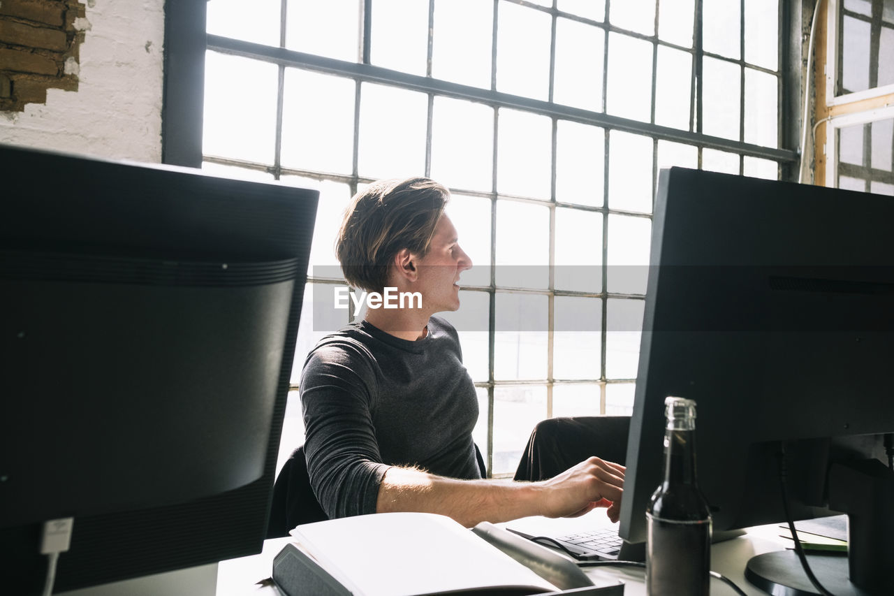 Young male hacker looking away while sitting at computer desk in creative workplace