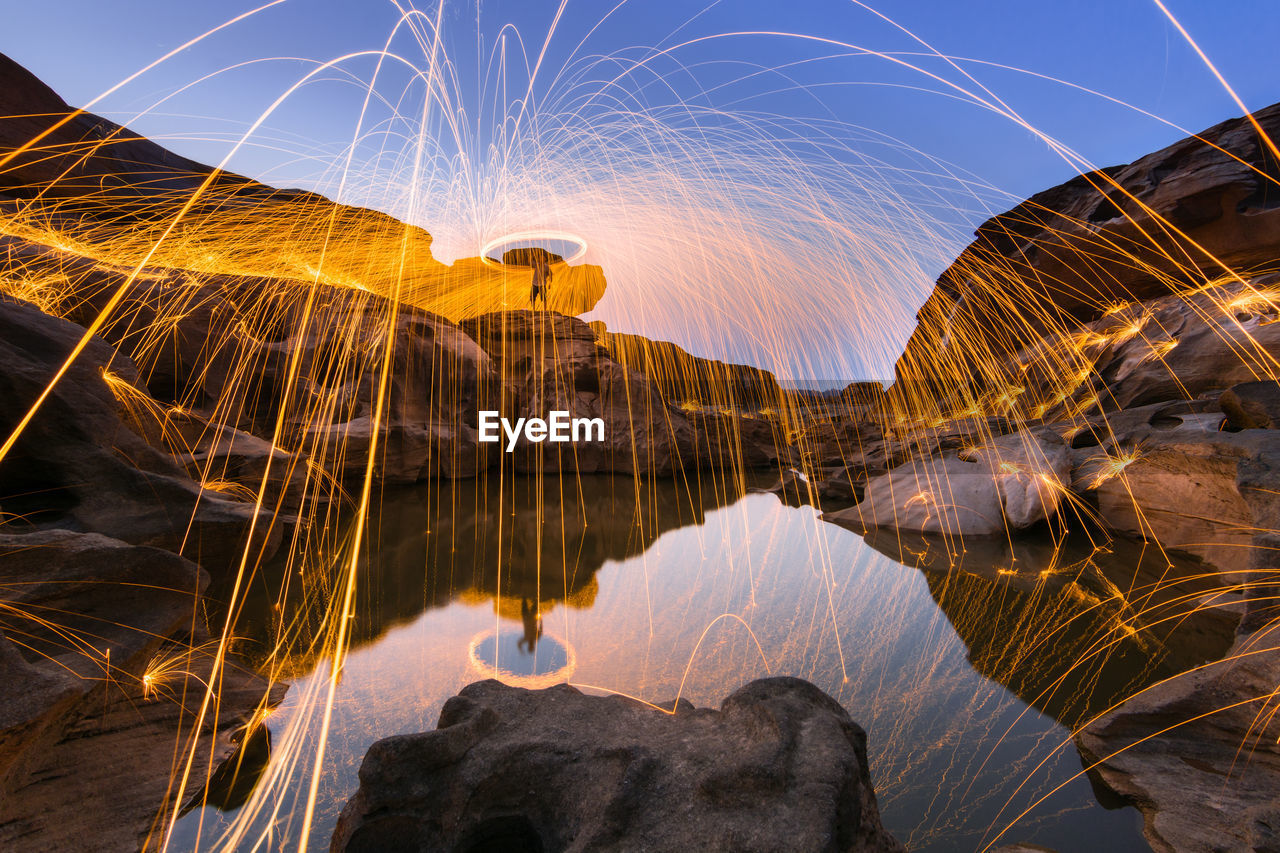 Light trails over lake on rock against sky at dusk