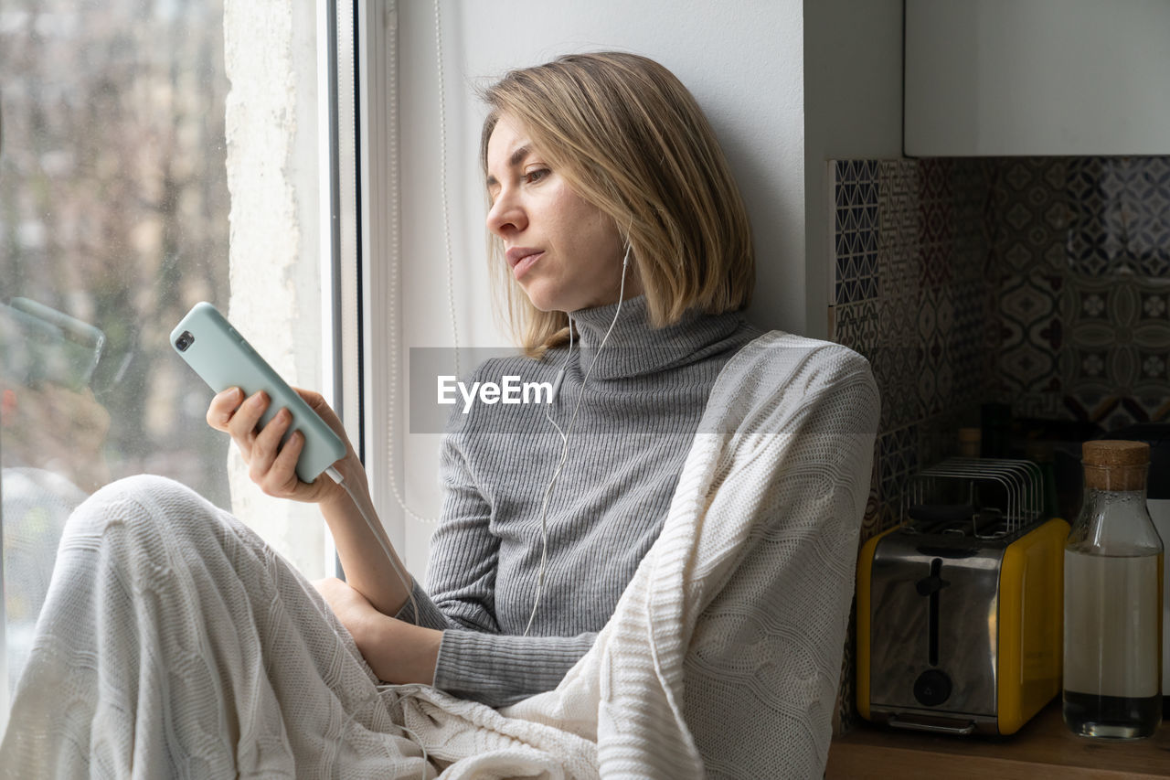 Bored female addicted and chatting in social media, sitting on windowsill at home using mobile phone