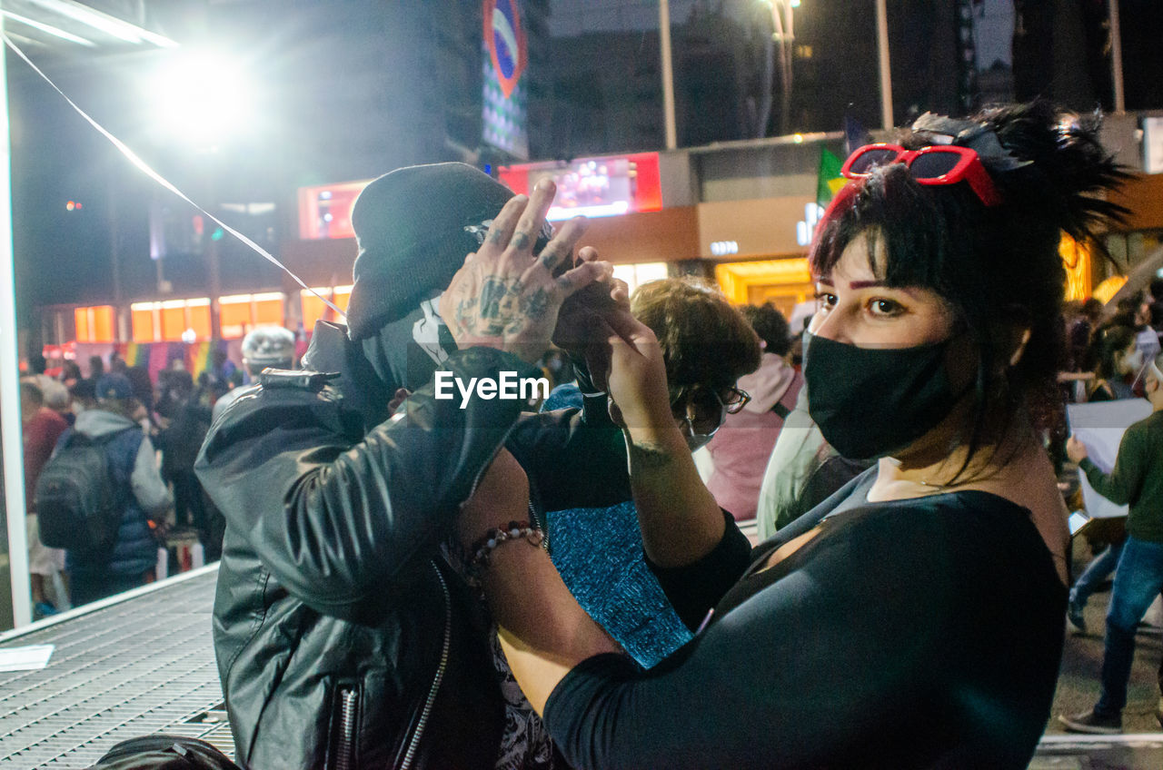 PORTRAIT OF WOMAN PHOTOGRAPHING AT ILLUMINATED STREET IN CITY