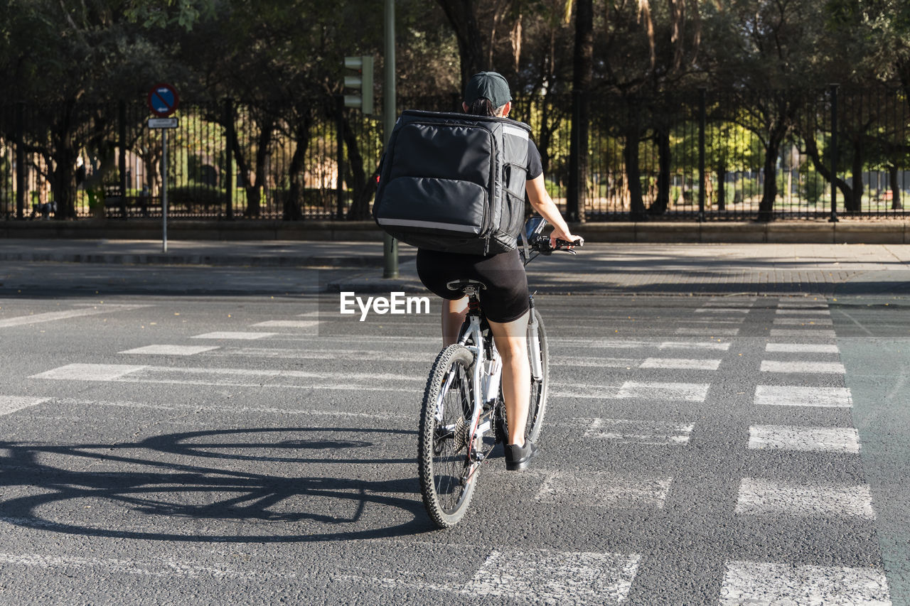 Young delivery woman with backpack riding bicycle on road during sunny day