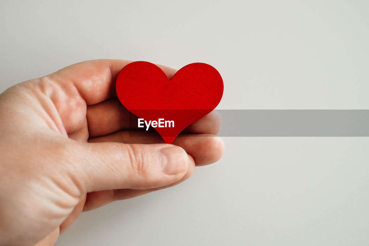 CLOSE-UP OF HAND HOLDING RED HEART SHAPE OVER WHITE BACKGROUND