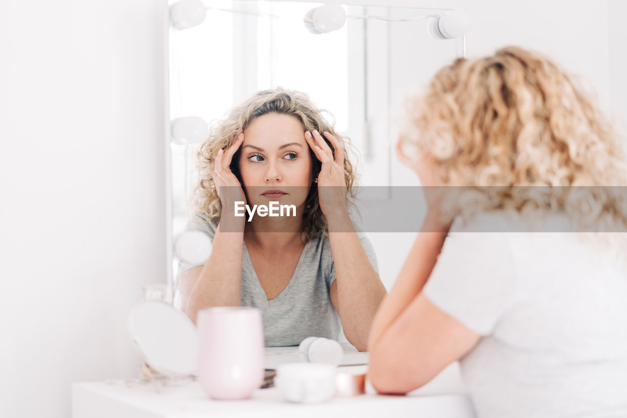 Unemotional attractive female in casual shirt touching face and looking at reflection in mirror attentively while sitting at vanity table in bedroom