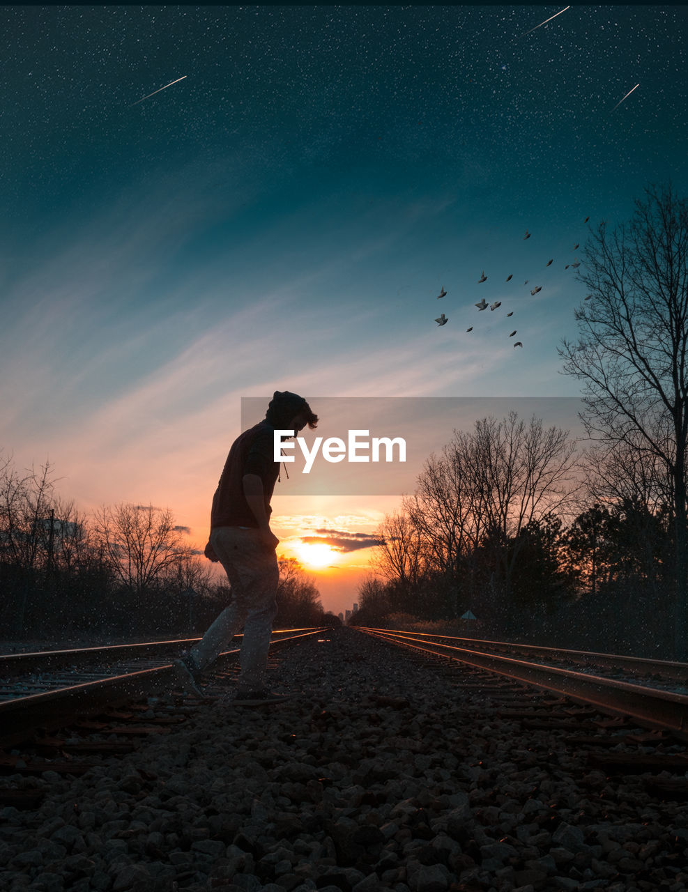 SILHOUETTE MAN STANDING ON RAILROAD TRACK AGAINST SKY AT SUNSET