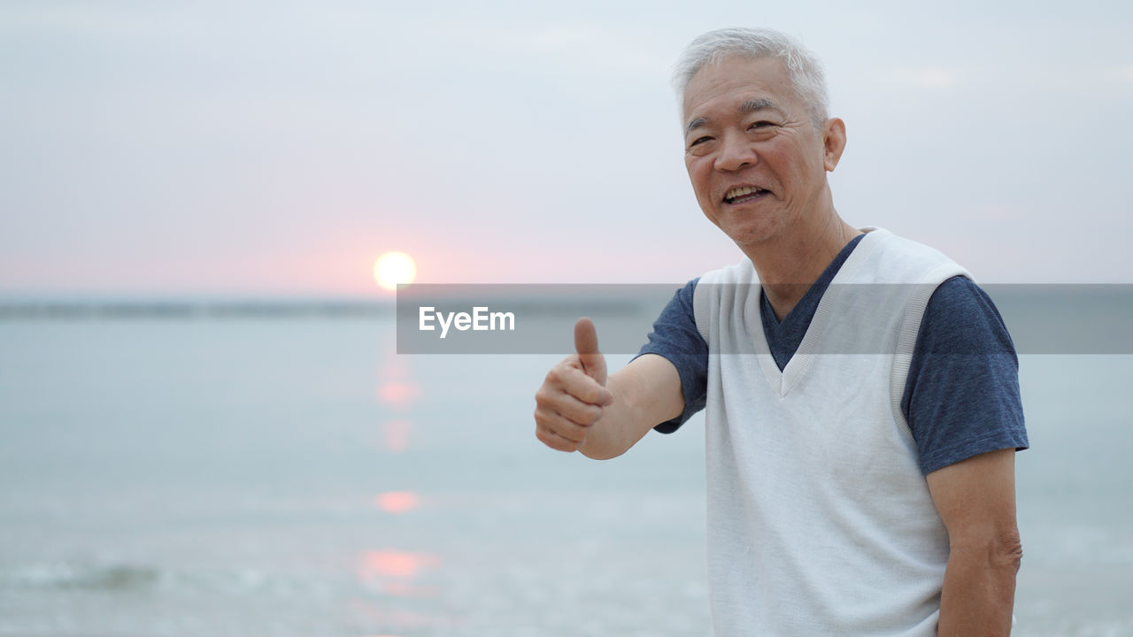Portrait of smiling man showing thumbs up while standing at beach during sunset