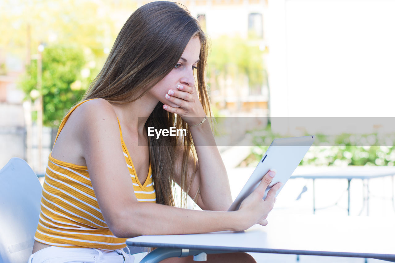 Shocked girl using mobile phone while sitting on table outdoors