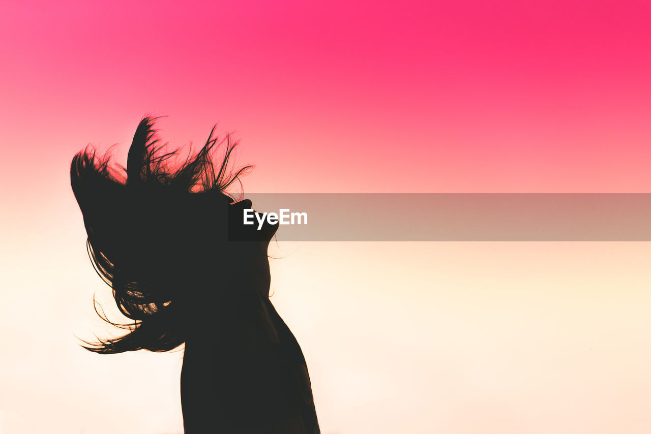 Side view of silhouette woman tossing hair against dramatic sky during sunset