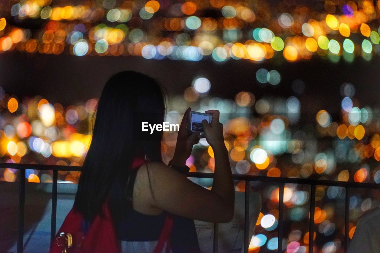 Woman photographing illuminated city through smart phone at night