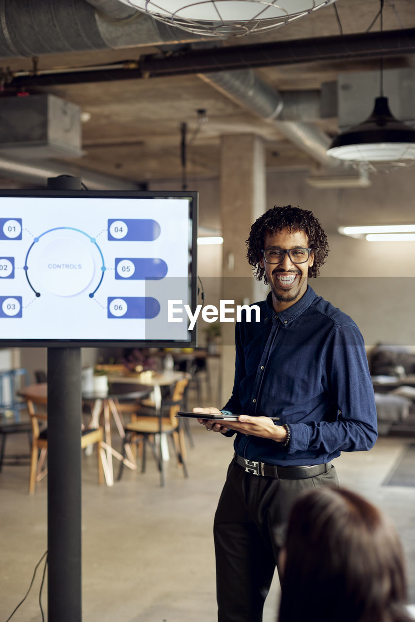 Happy businessman holding digital tablet while standing by television set during meeting in office