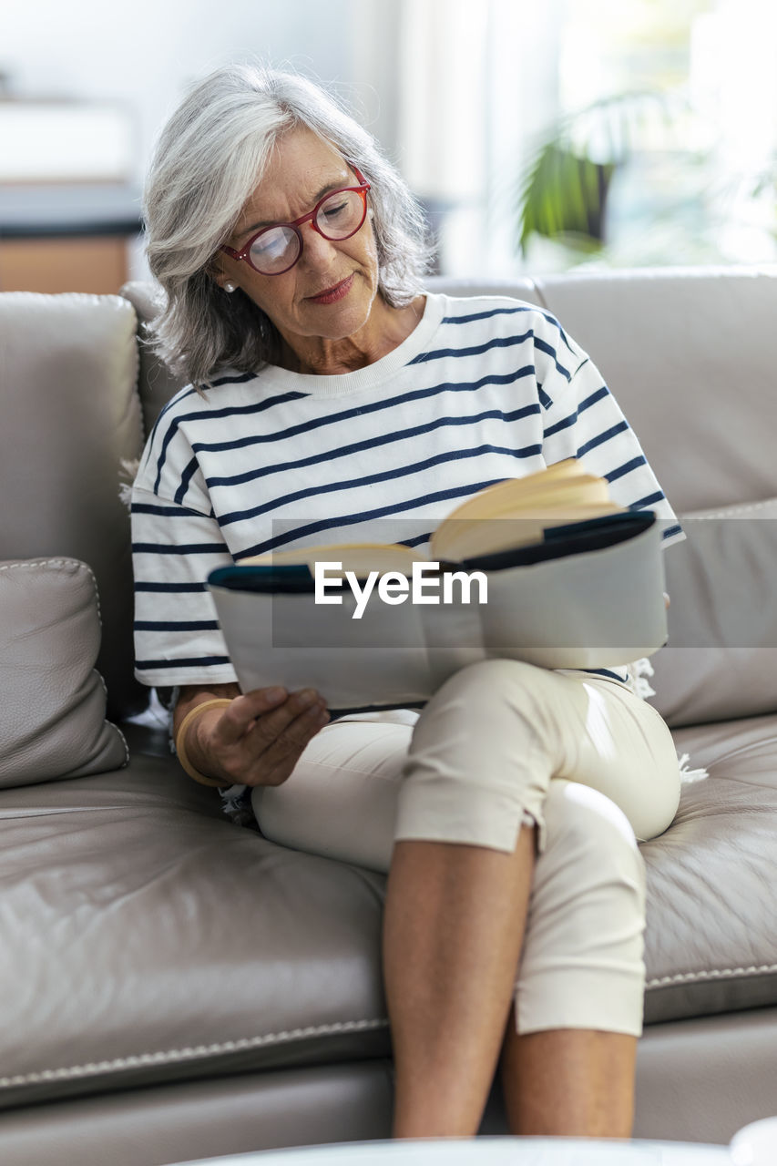 Woman reading book sitting with legs crossed on sofa at home