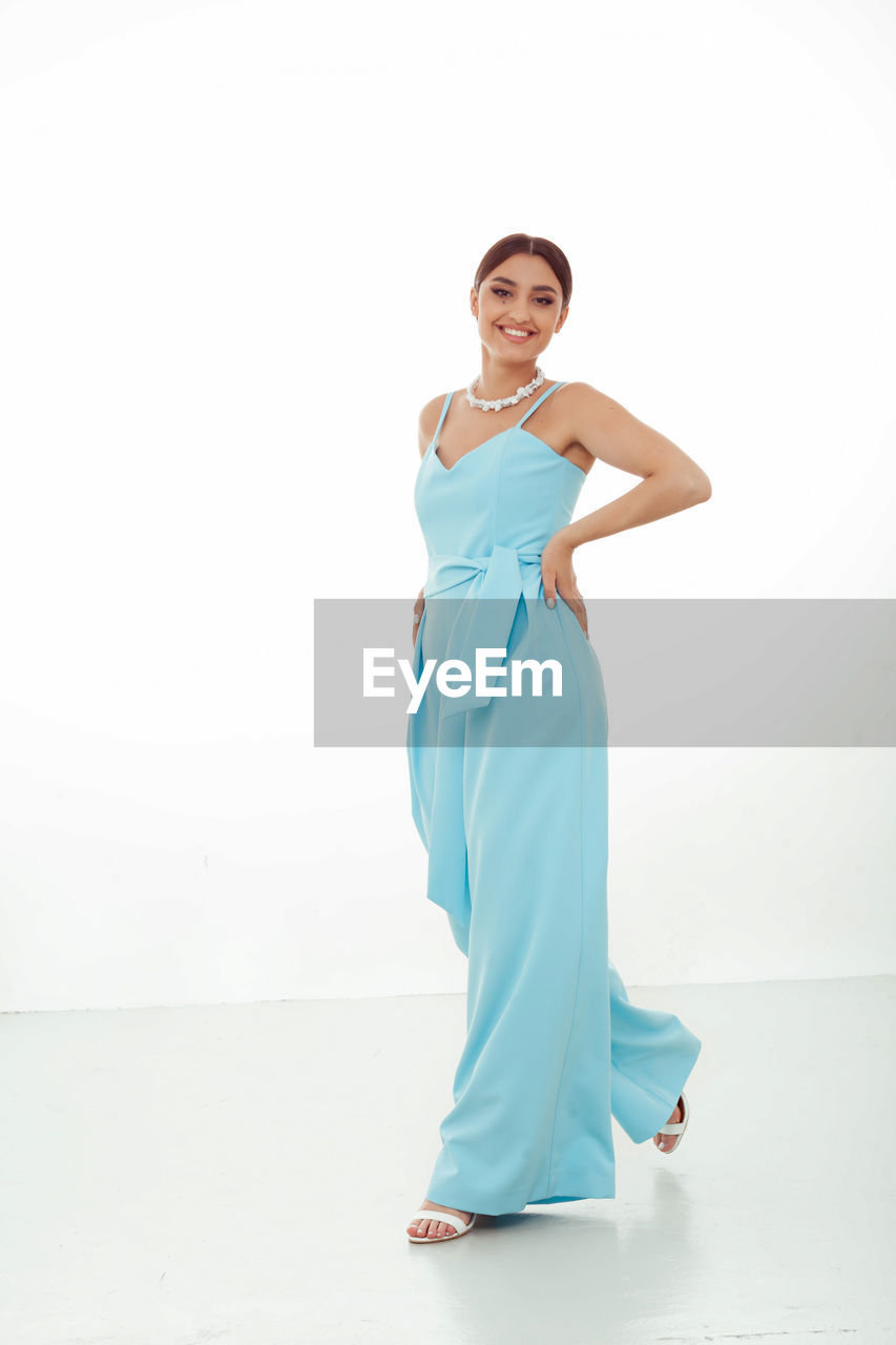 full length, one person, adult, portrait, women, gown, looking at camera, studio shot, clothing, young adult, indoors, standing, dress, smiling, fashion, white background, female, bridal clothing, happiness, copy space, front view, emotion, wedding dress, elegance, hand on hip, lifestyles, hand, brown hair
