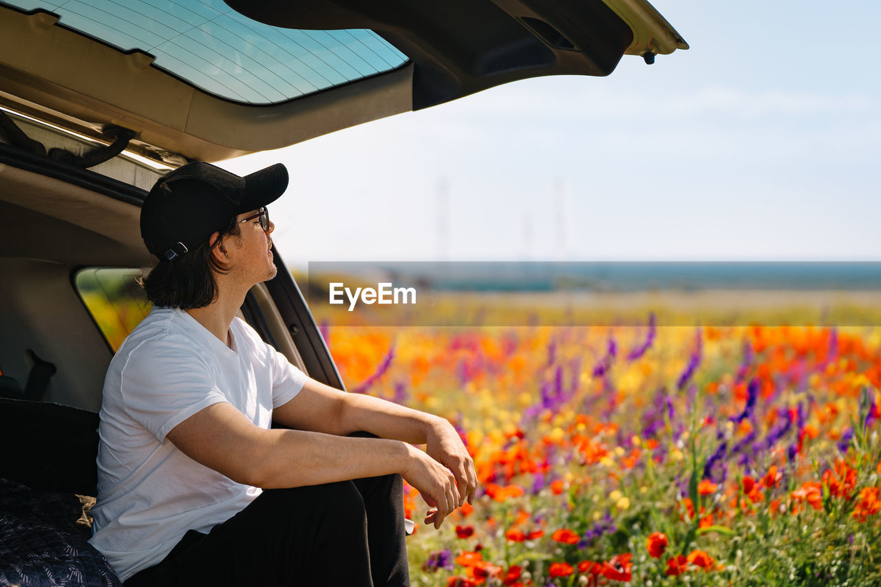 Side view of young man standing in suv car  by flowering wild plants in summer