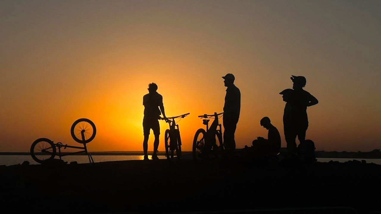 sunset, silhouette, sky, group of people, bicycle, men, orange color, nature, evening, horizon, adult, dusk, transportation, lifestyles, sun, leisure activity, activity, beach, sea, water, standing, copy space, outdoors, small group of people, travel, sunlight, beauty in nature, full length, land