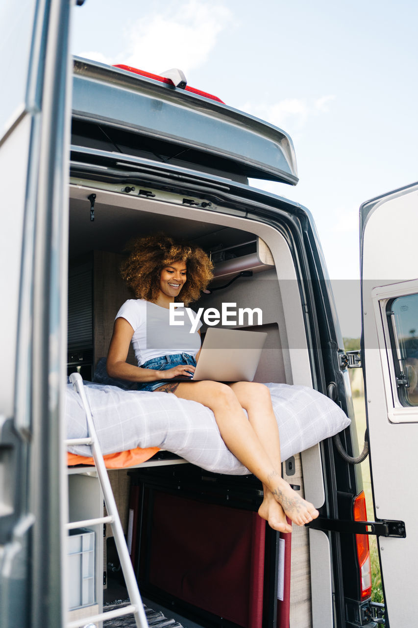 Young african american female traveler with curly hair watching movie on laptop while resting inside camper van during summer holidays