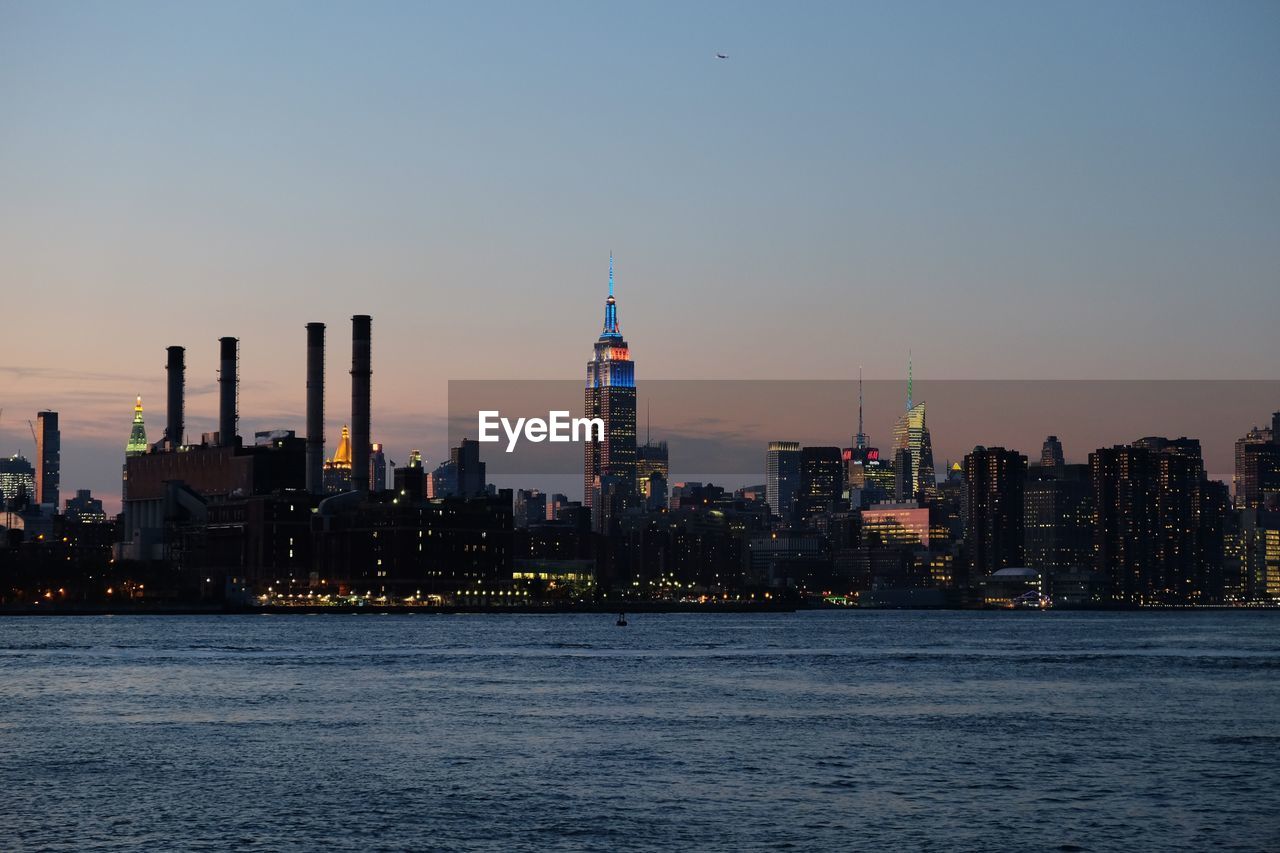 East river by illuminated empire state building in city against sky during sunset