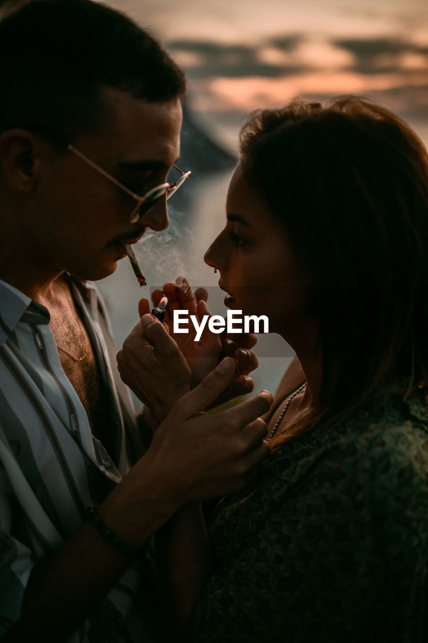 Man lighting cigarette for girlfriend while chilling on seashore at sunset